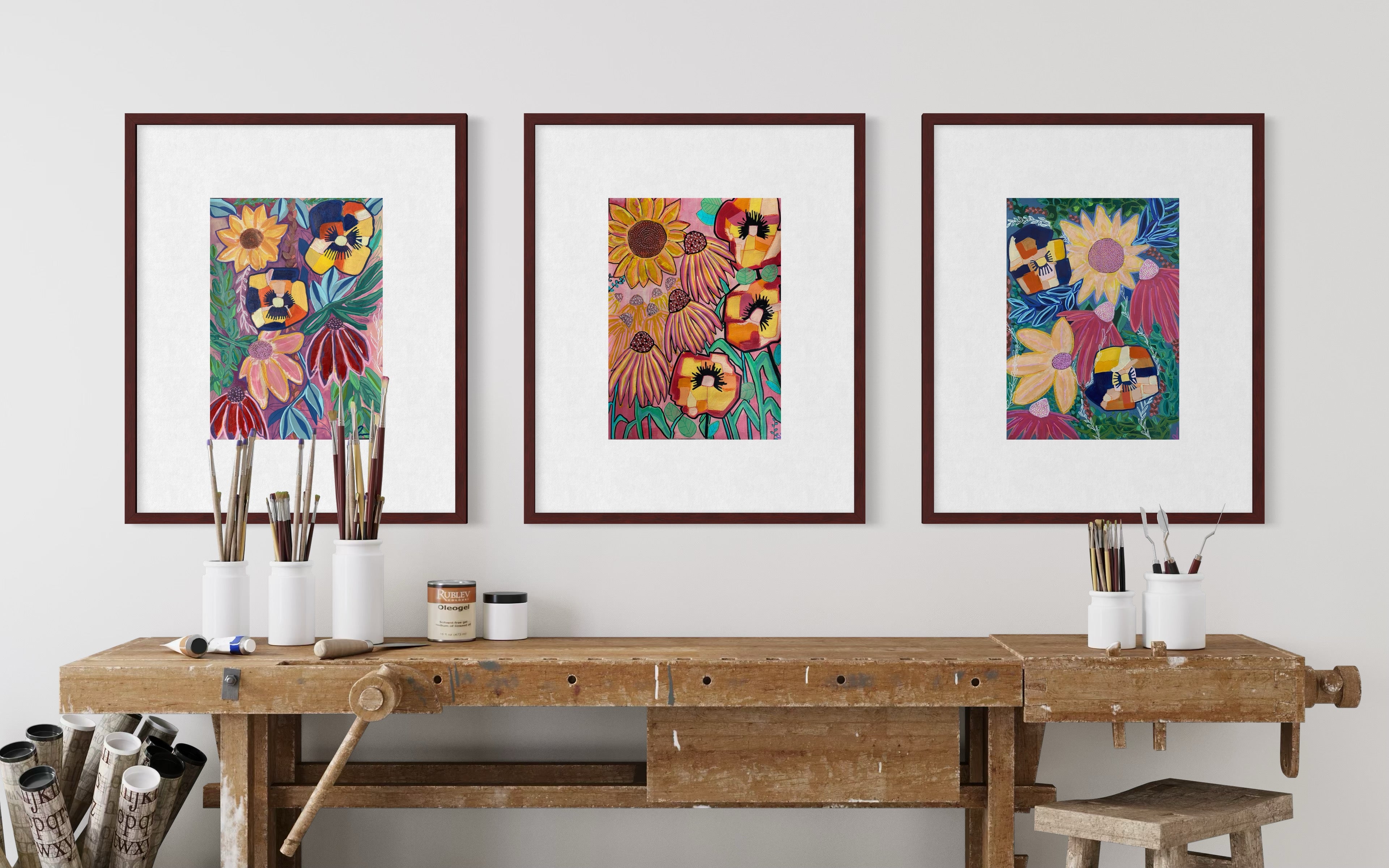 Three framed colorful floral art prints hang on a light gray wall above a rustic wooden workbench with paintbrushes and art supplies.