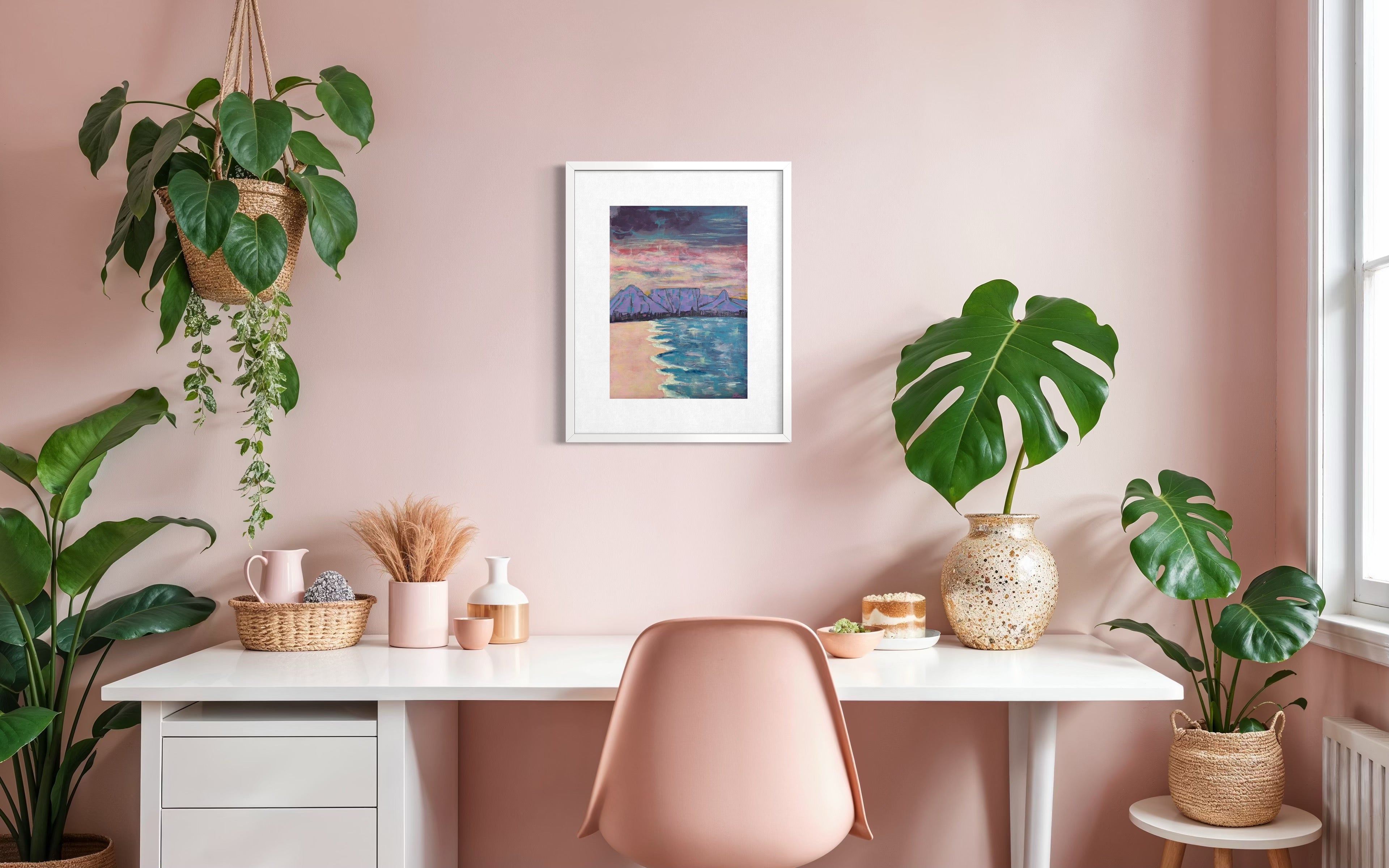 A framed art print depicting a beach sunset hangs on a dusty pink wall above a white desk with a blush pink chair, surrounded by potted green plants in a bright, modern home office setting.