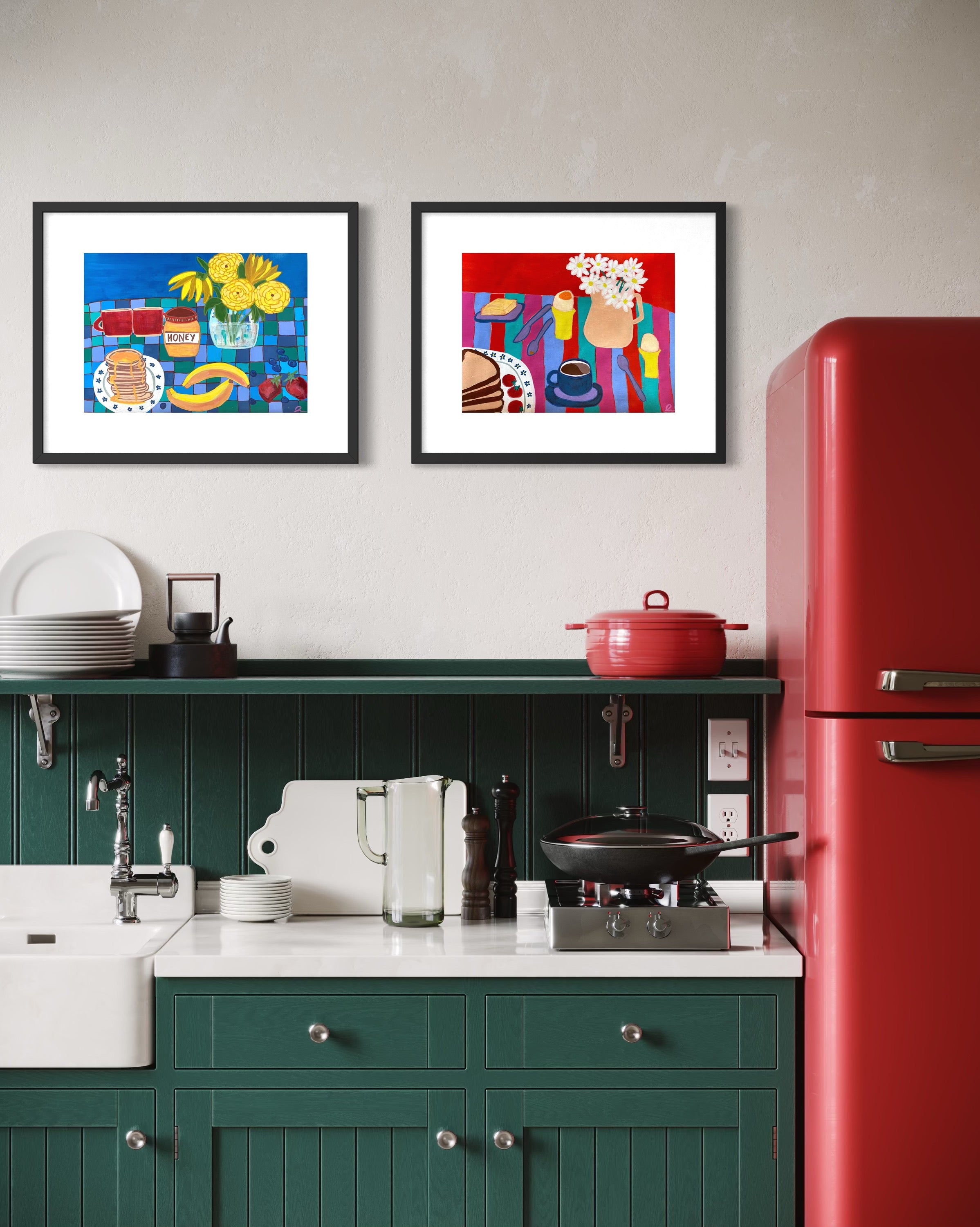 Two colorful framed art prints hang on a light gray wall above a dark green kitchen counter with a white sink, red refrigerator, and kitchen accessories.