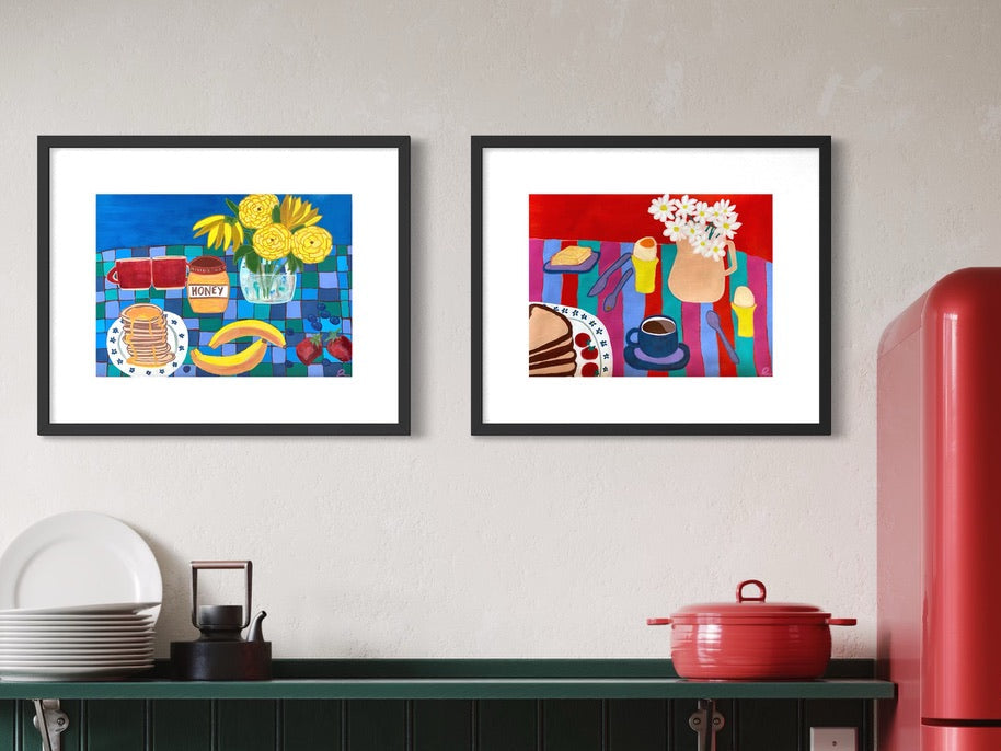 Two framed colorful still life paintings hang on a light beige wall above a dark green kitchen counter with a red pot and a red refrigerator.